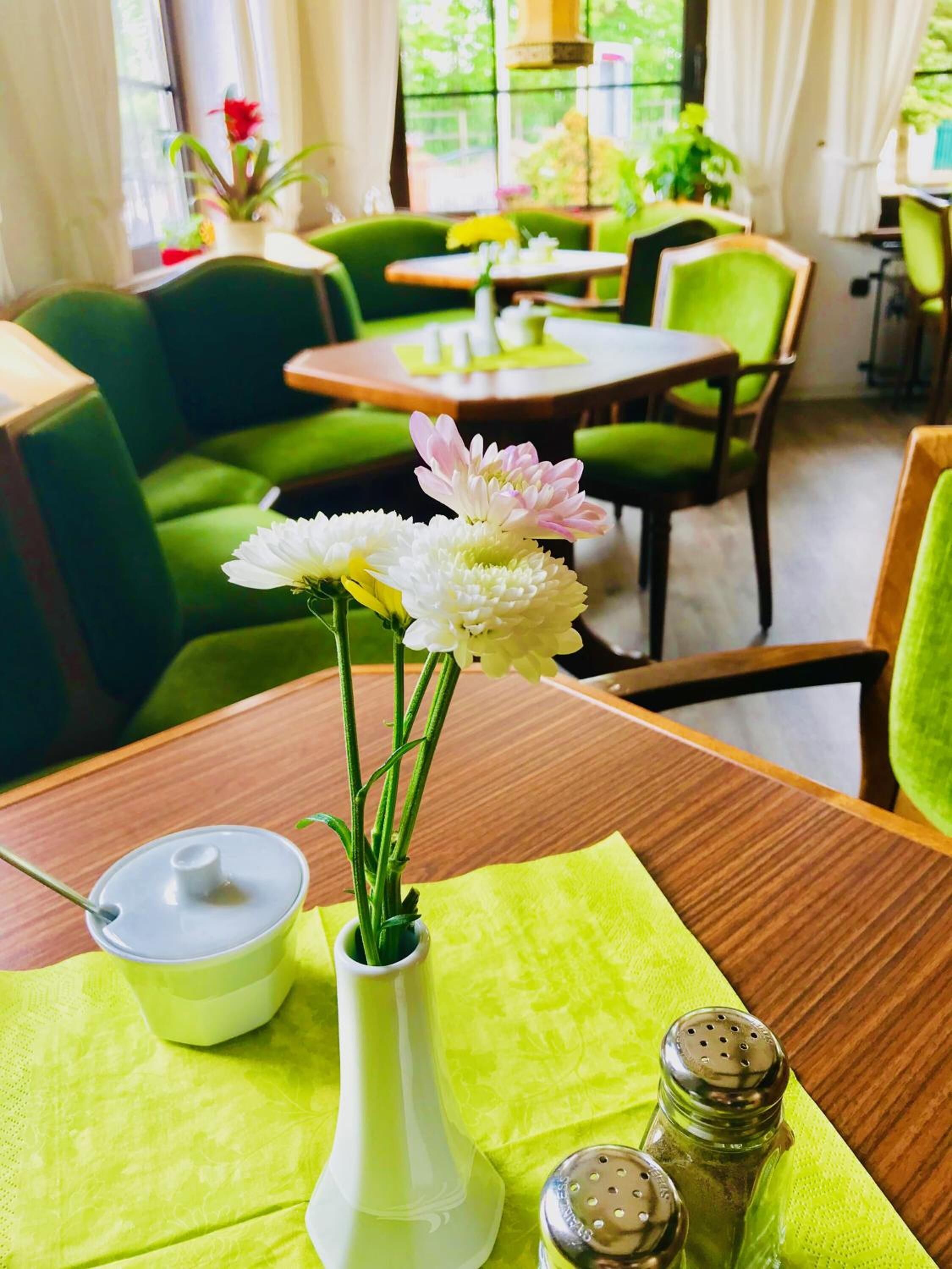 A dining area with green upholstered chairs, a wooden table with a white vase containing flowers, a green napkin, and salt and pepper shakers.