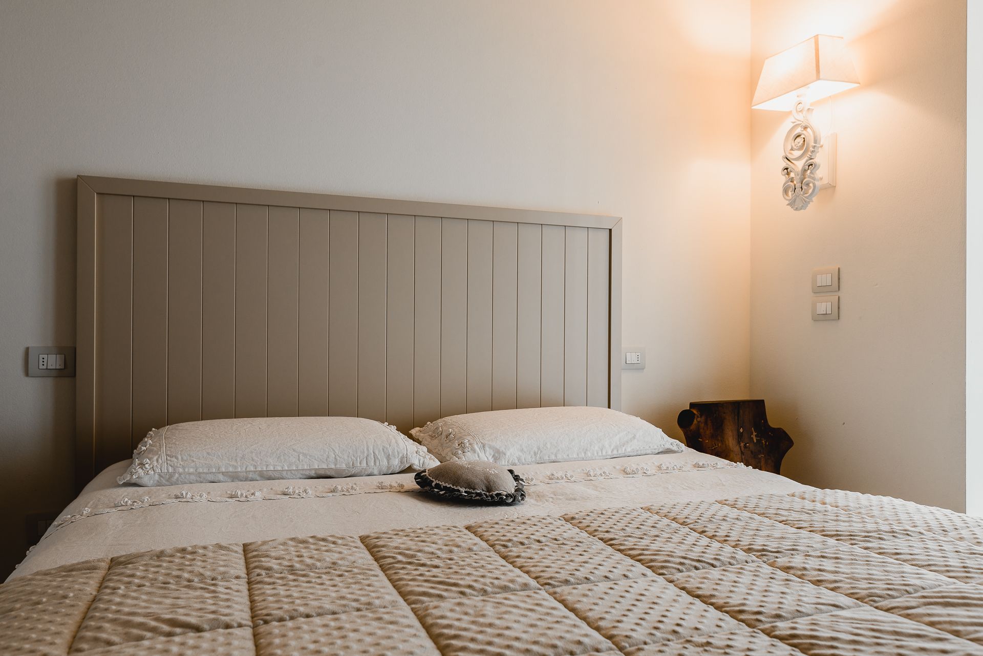 A neatly made bed with a quilted cover, a wooden headboard, and a wall-mounted light fixture.
