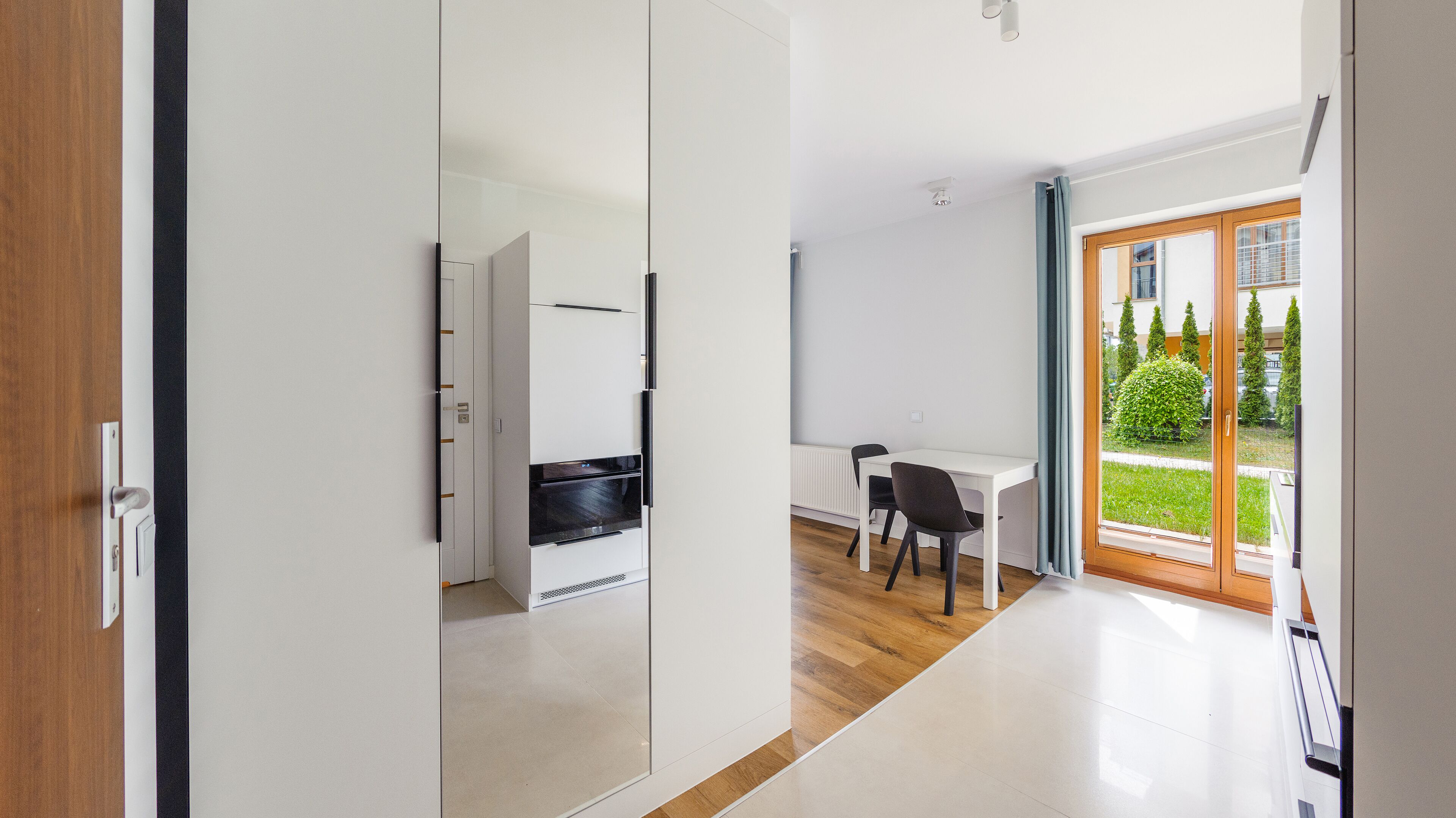 A modern kitchen with white cabinets and a black oven, a dining area with a white table and black chairs, and a large sliding glass door leading to a garden.