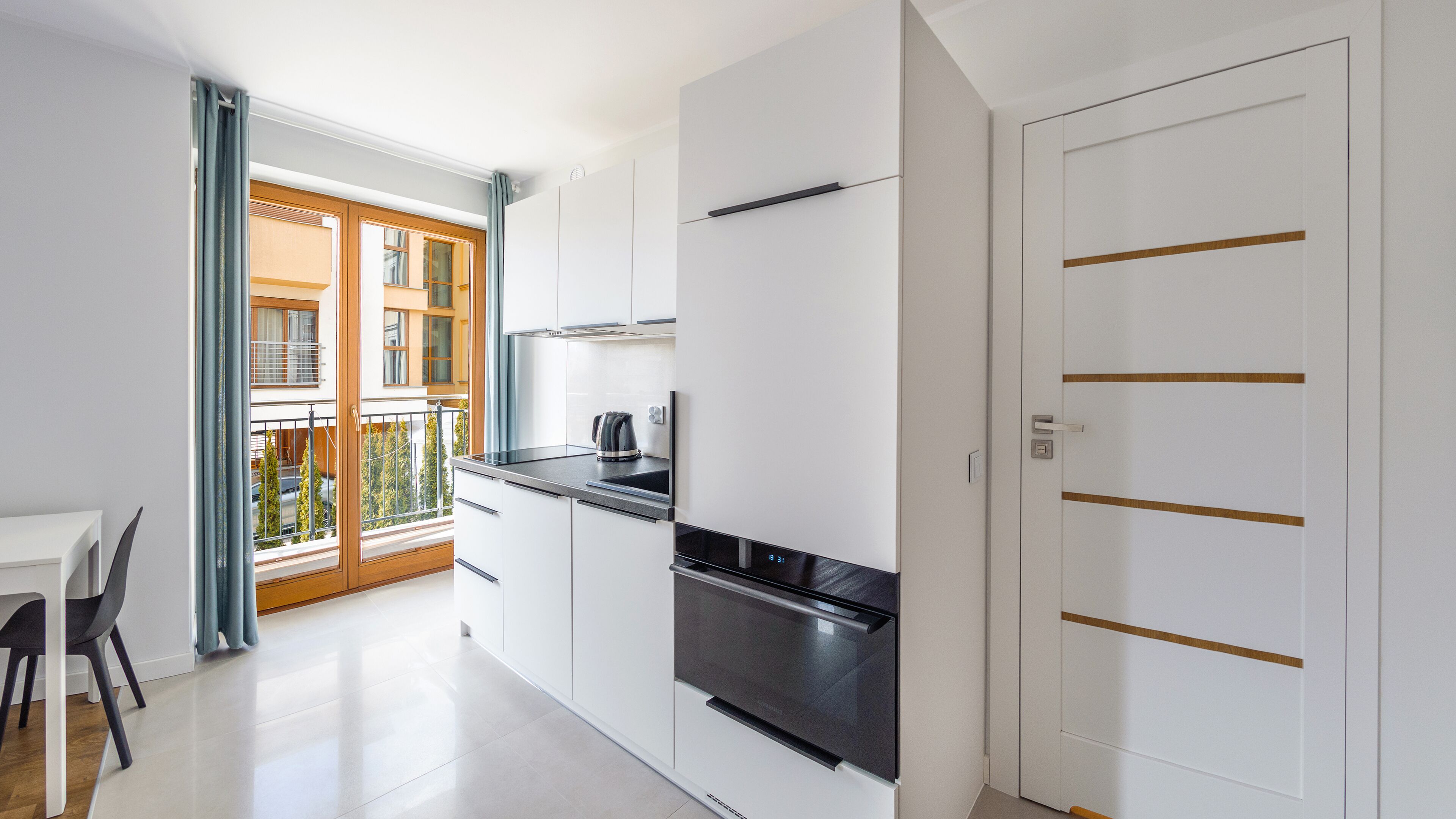 A modern kitchen with white cabinets, a black oven, and a view of a balcony with wooden doors and a railing.