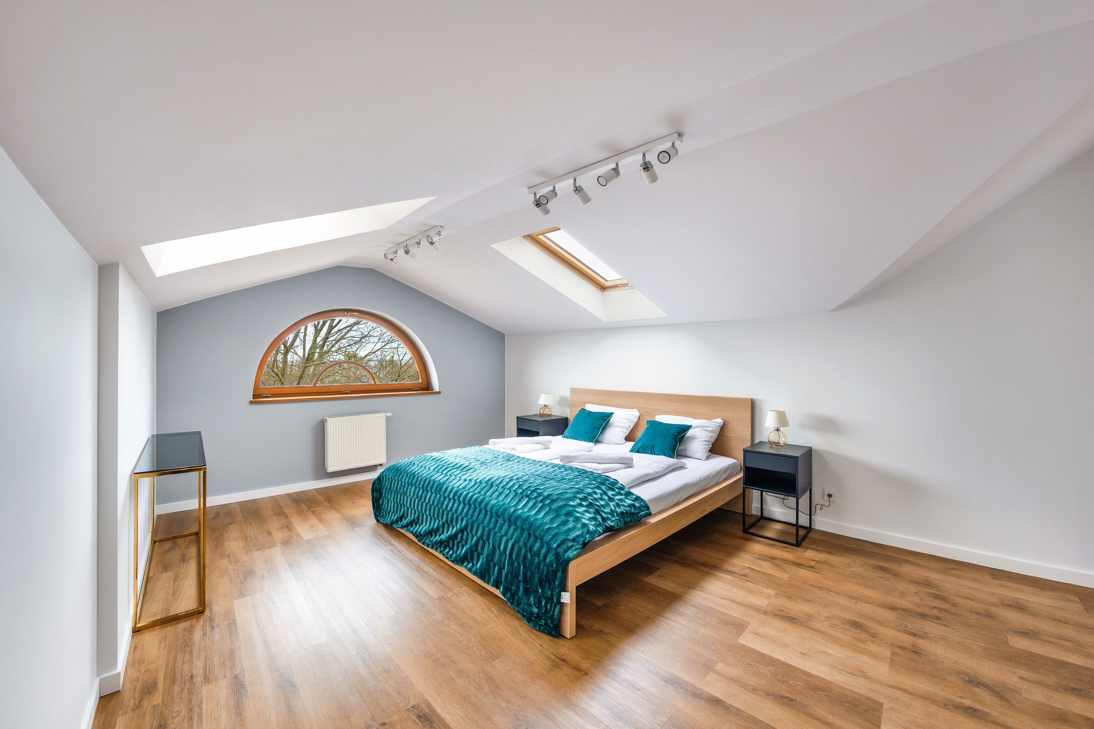 Un dormitorio con tragaluz, una cama con sábanas blancas, una manta verde y una mesita de noche con lámpara.