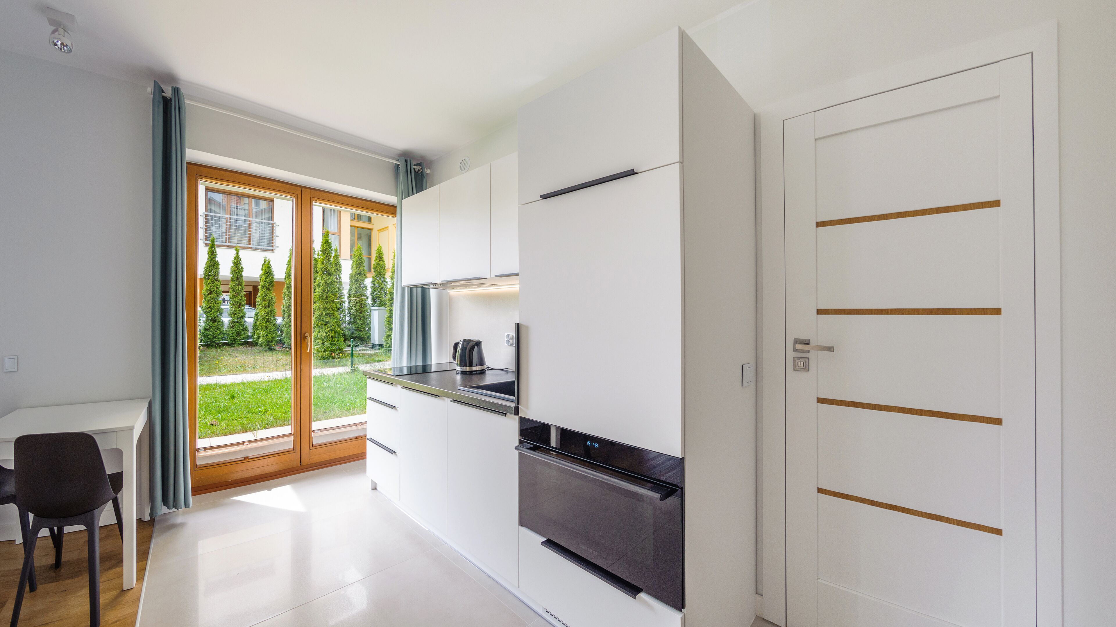 A modern kitchen with white cabinets, a black countertop, and a built-in oven. There is a sliding glass door leading to a balcony with a view of trees and a building.