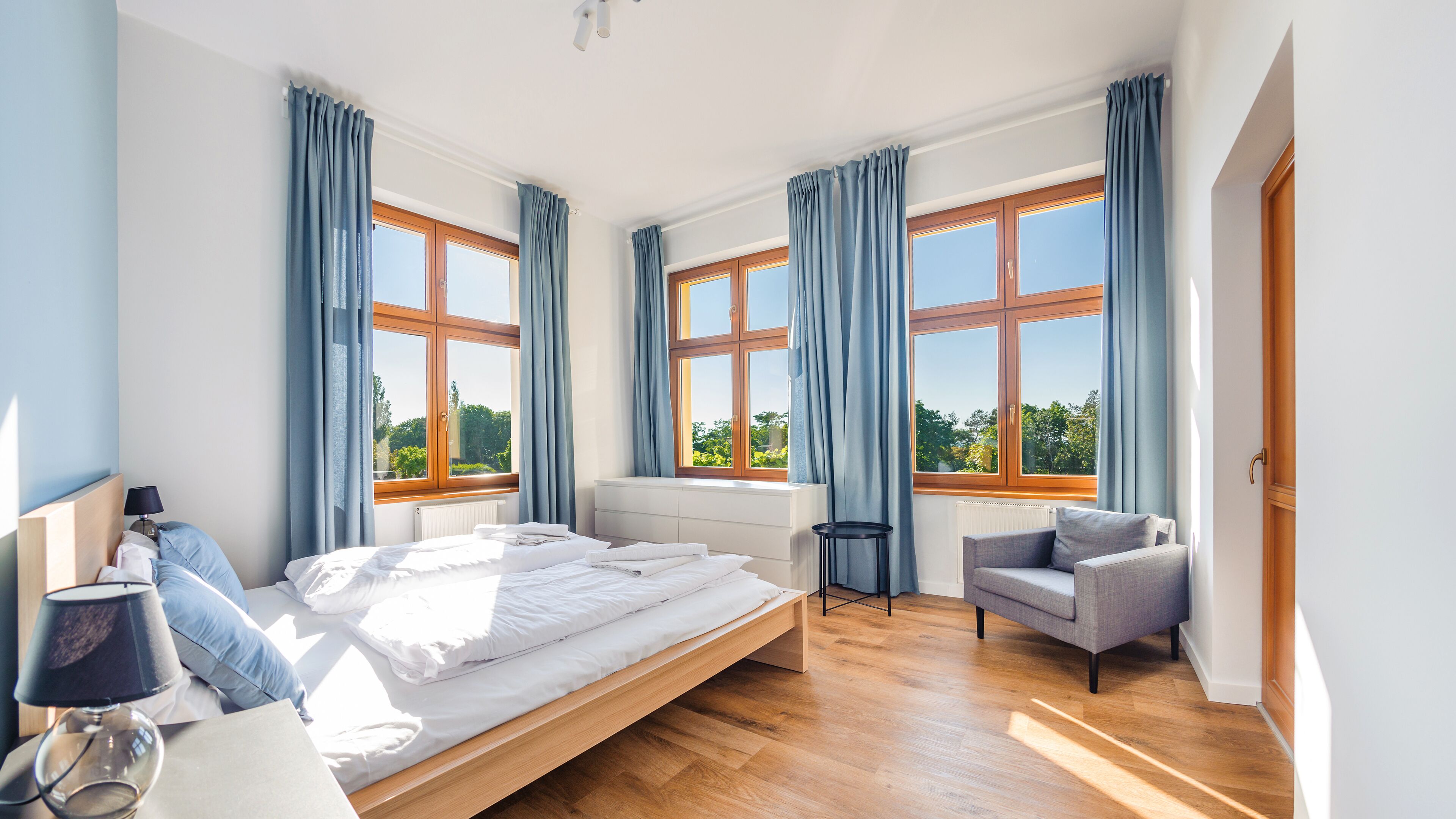 A modern bedroom with a wooden bed, a grey armchair, and large windows with blue curtains.
