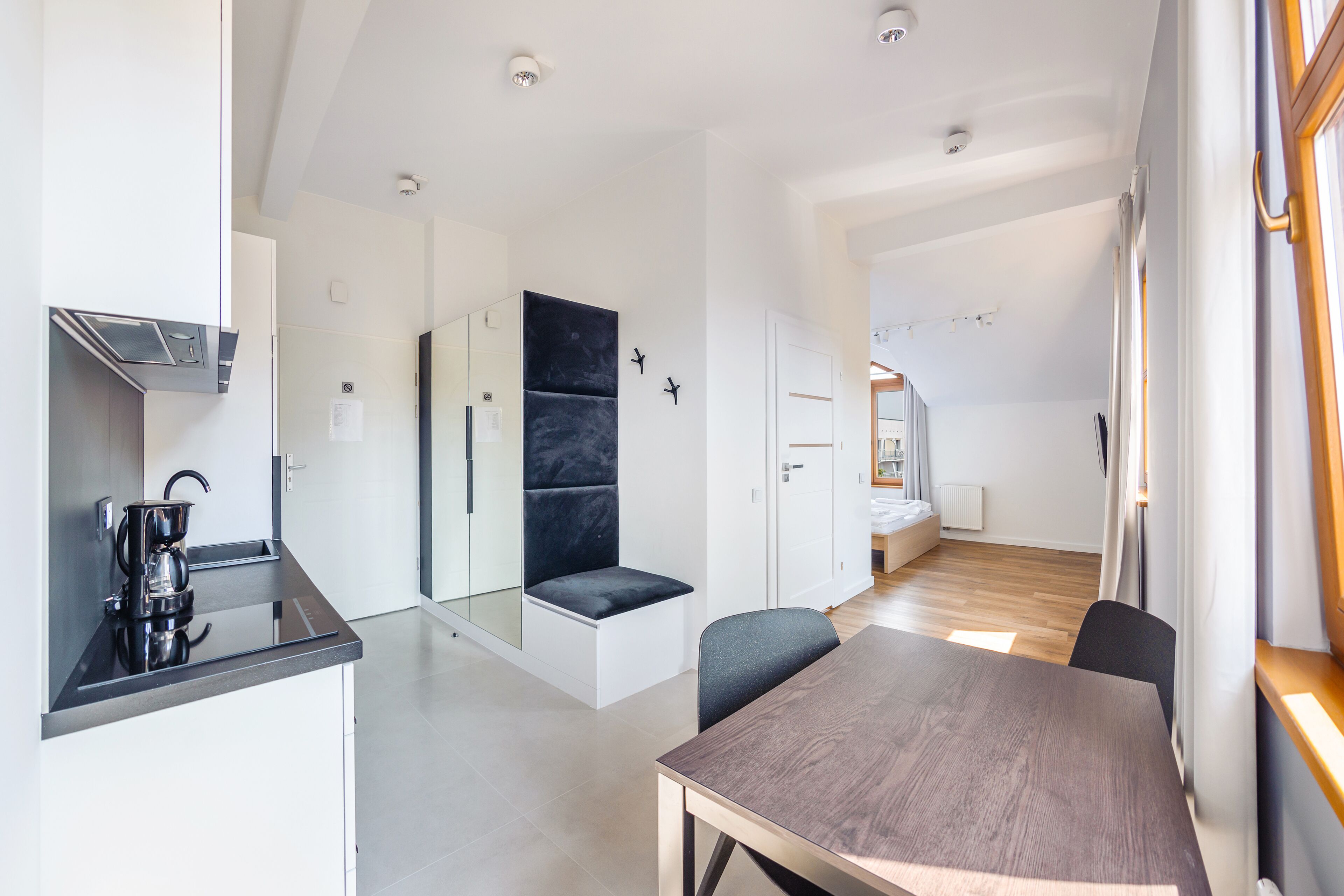 Een moderne keuken met een koffiezetapparaat, een zithoek met een comfortabele bank, een eettafel met stoelen, en uitzicht op een slaapkamer met een bed en een raam.