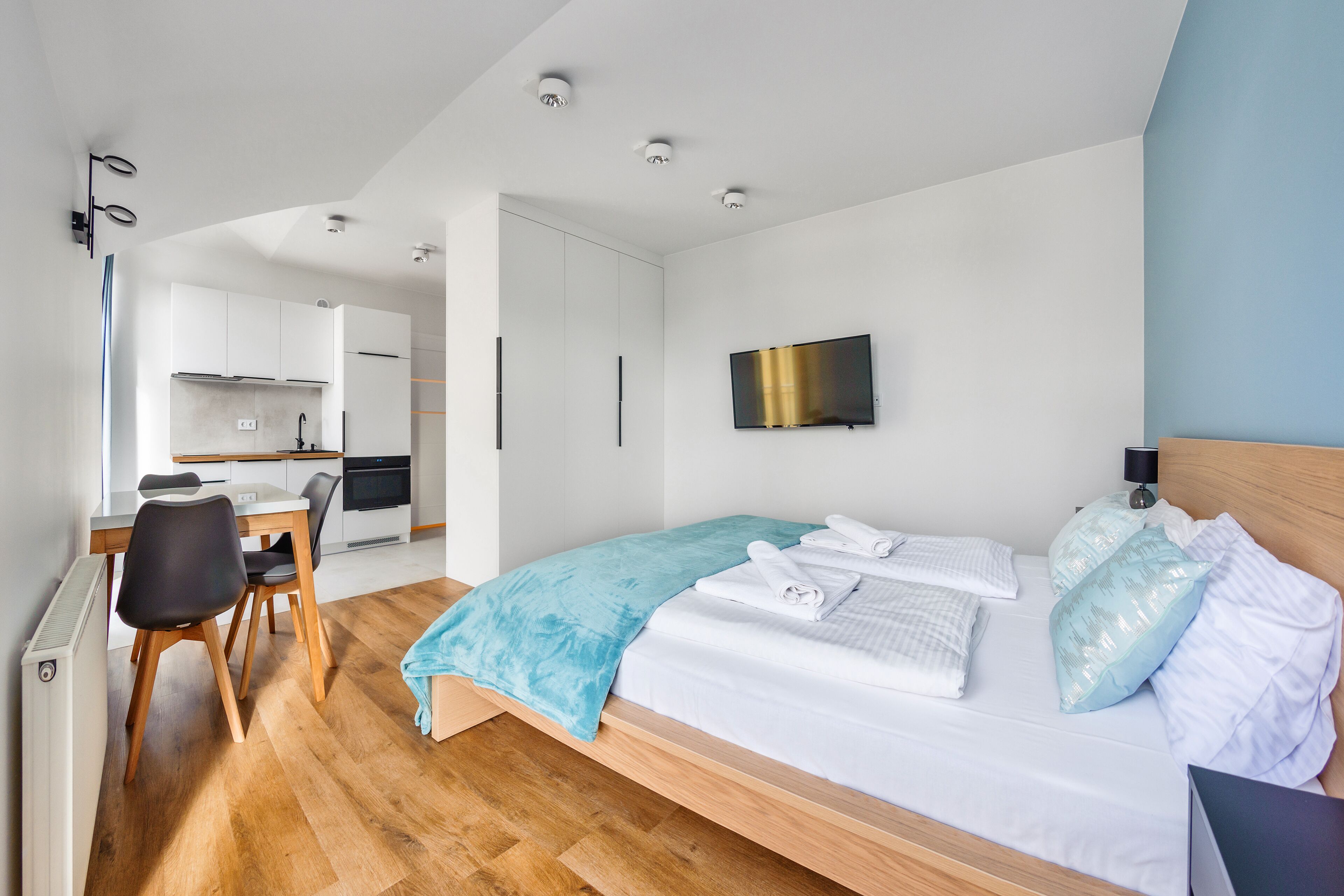 Una habitación de hotel moderna con piso de madera, una cama con sábanas blancas, una manta azul y un televisor de pantalla plana en la pared.