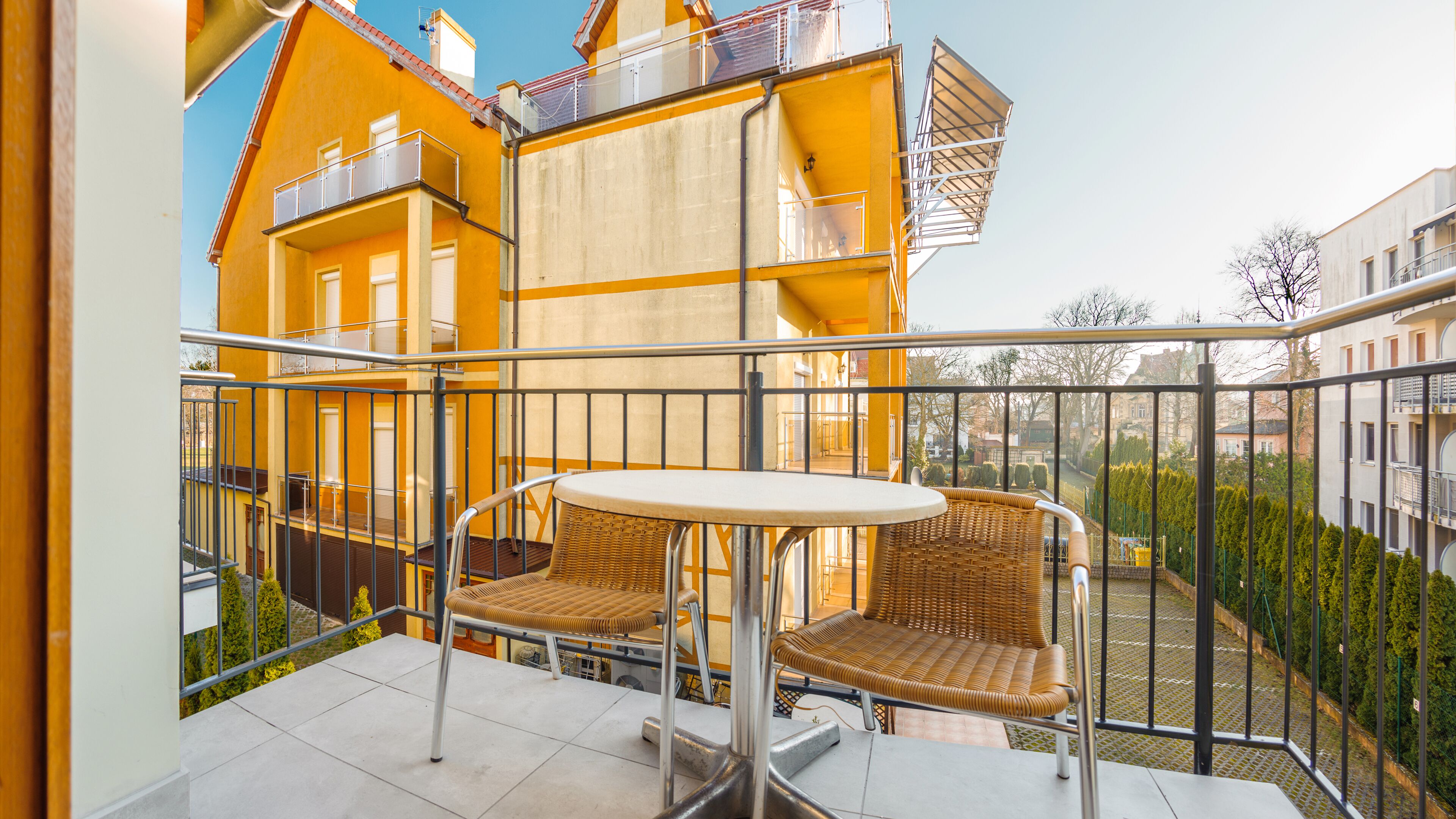 A balcony with wicker chairs and a table, overlooking a courtyard with buildings and trees.