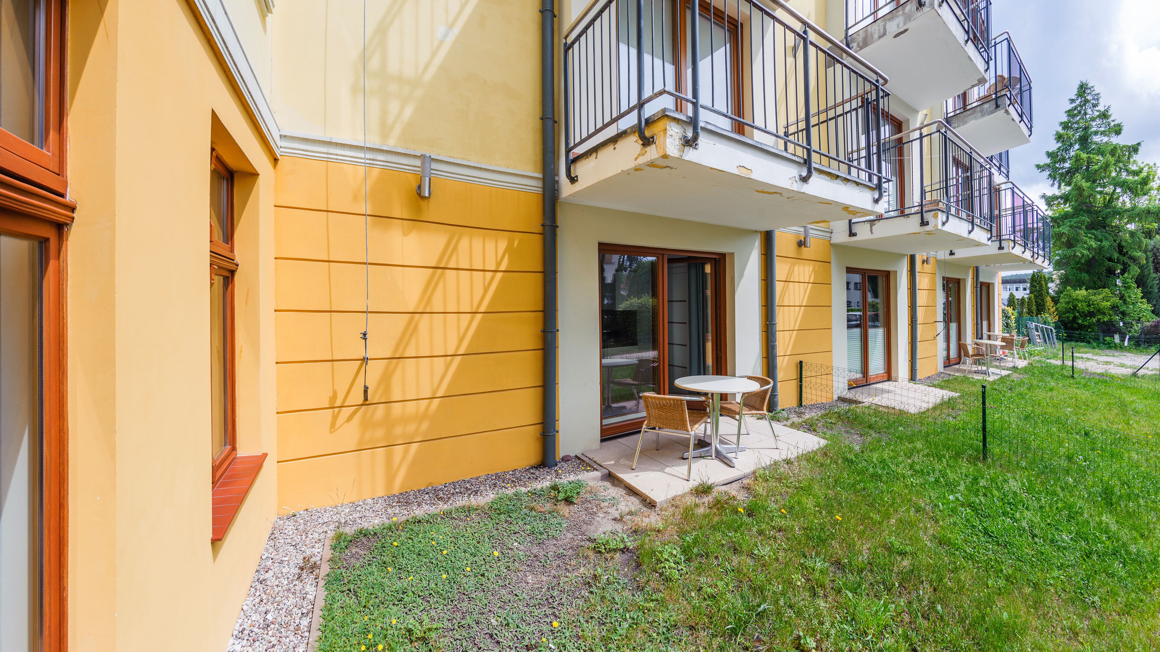 A yellow building with a balcony, a small patio with a table and chairs, and a grassy area.