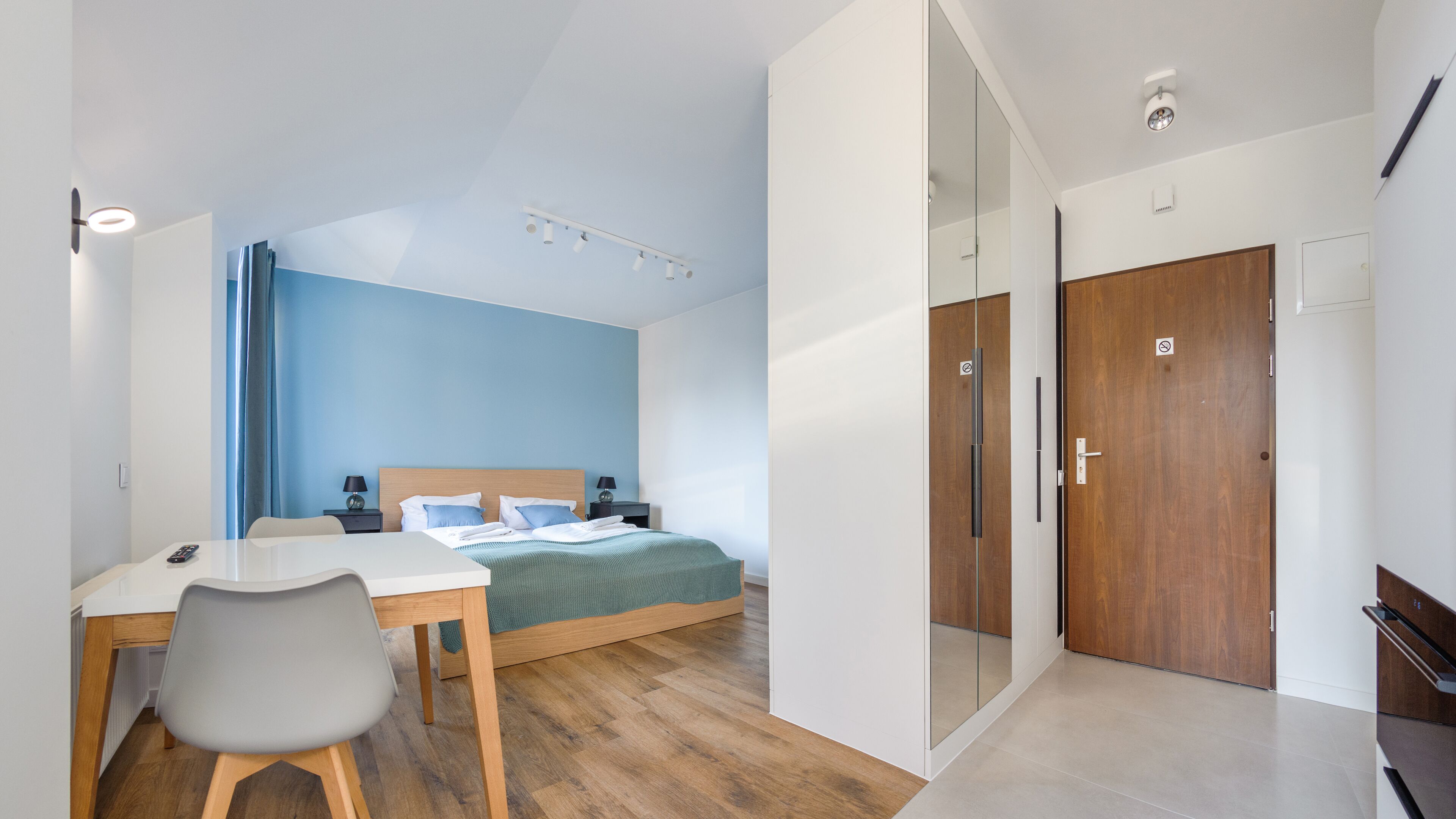 A modern hotel room with a bed, a desk, and a chair. There is a wooden door and a glass partition.