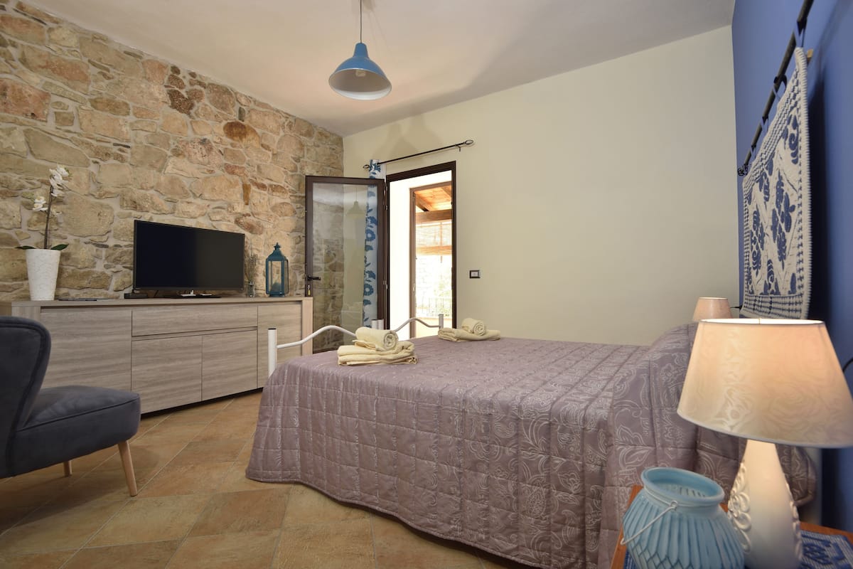 Une chambre avec un mur en pierre, un lit recouvert d’une couverture violette, un téléviseur posé sur un meuble en bois, une chaise et une lampe.