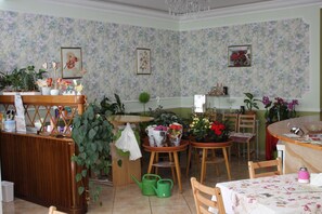 A cozy, well-lit room with a wooden reception desk, several chairs, and a dining table. There are potted plants and flowers on the walls and tables.