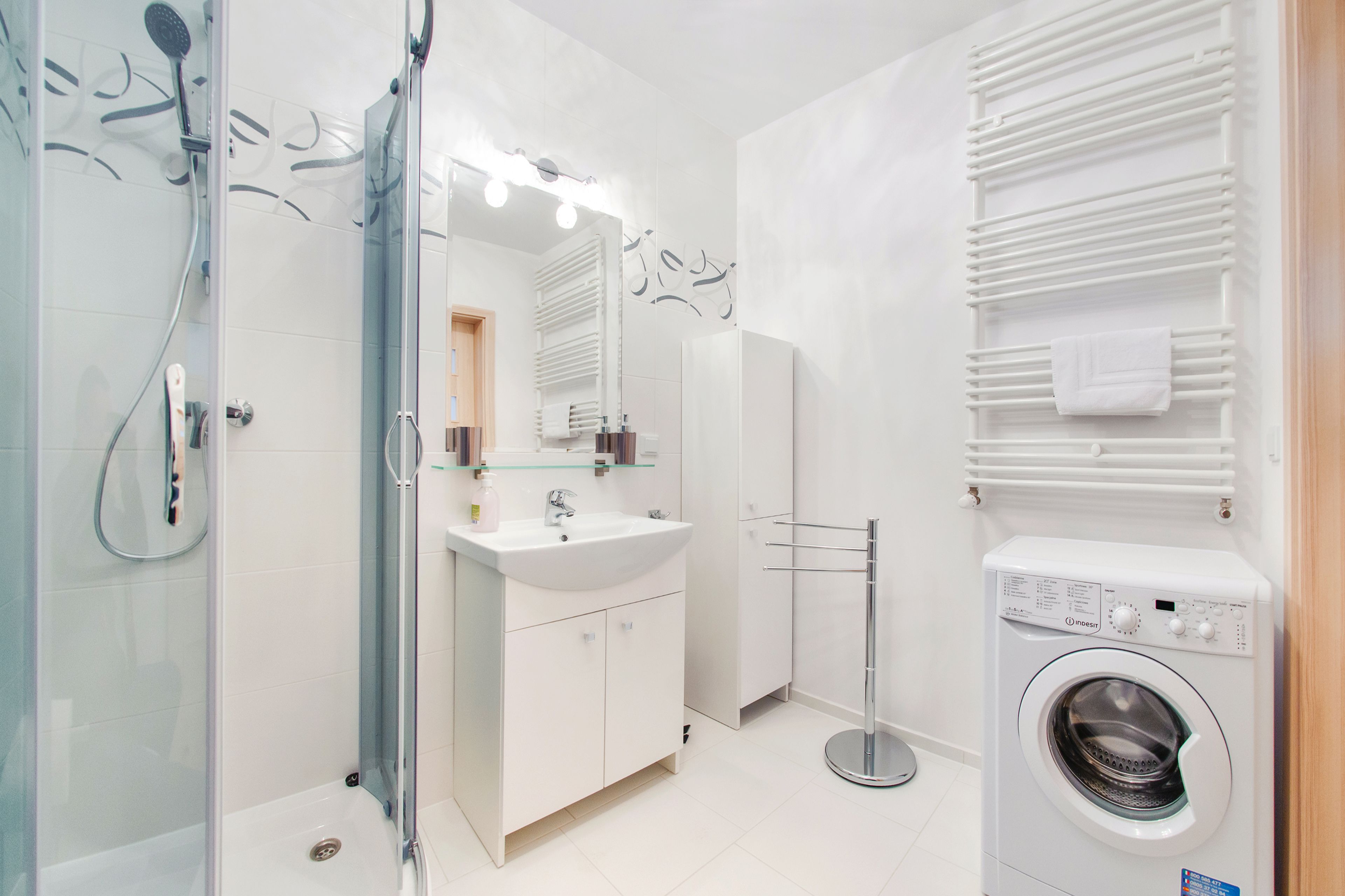 A modern bathroom with a glass-enclosed shower, a white sink, a mirror with lights, and a built-in washing machine.
