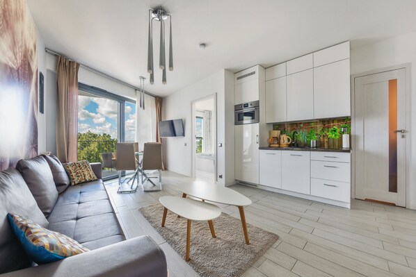 Ein modernes Wohnzimmer mit Sofa, Couchtisch und Essbereich. Es beinhaltet eine Küche mit weißen Schränken und einem eingebauten Backofen.
