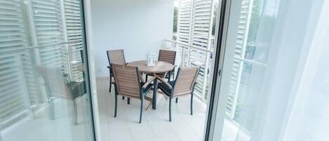 Un balcón con mesa y sillas, ventanas amplias con persianas y vistas al exterior.