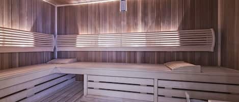 Sauna, jossa on puiset penkit ja säleikköseinät. Siinä on penkki, jossa on sisäänrakennettu istuinosa, sekä pieni ämpäri.