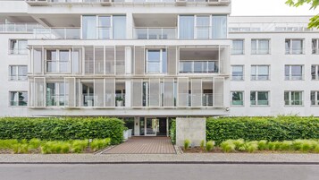A modern multi-story building with balconies, a gated entrance, and a well-maintained hedge.
