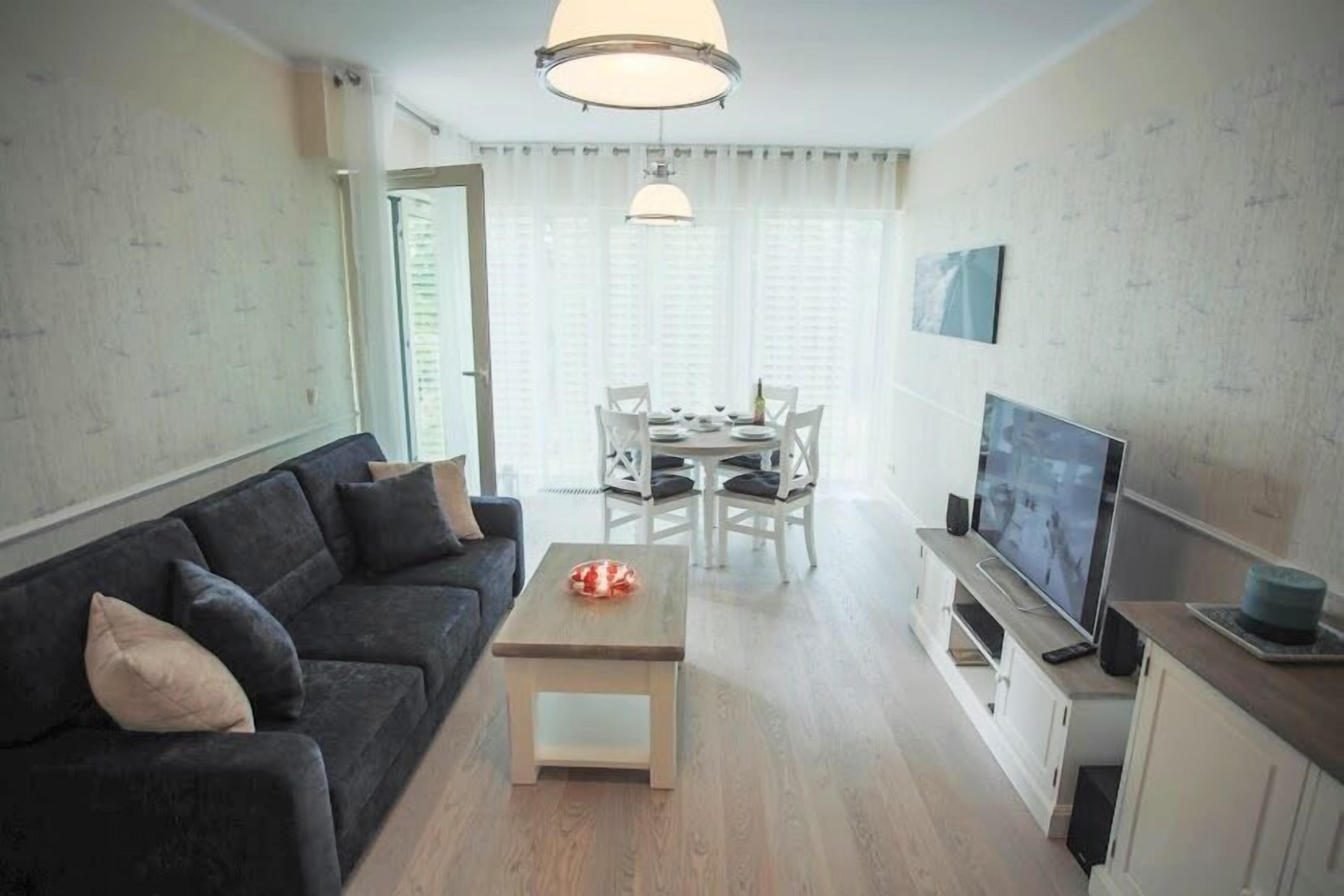 Una sala moderna con un sofá oscuro, una pequeña mesa de centro de madera, un comedor con sillas blancas y un televisor sobre un mueble.