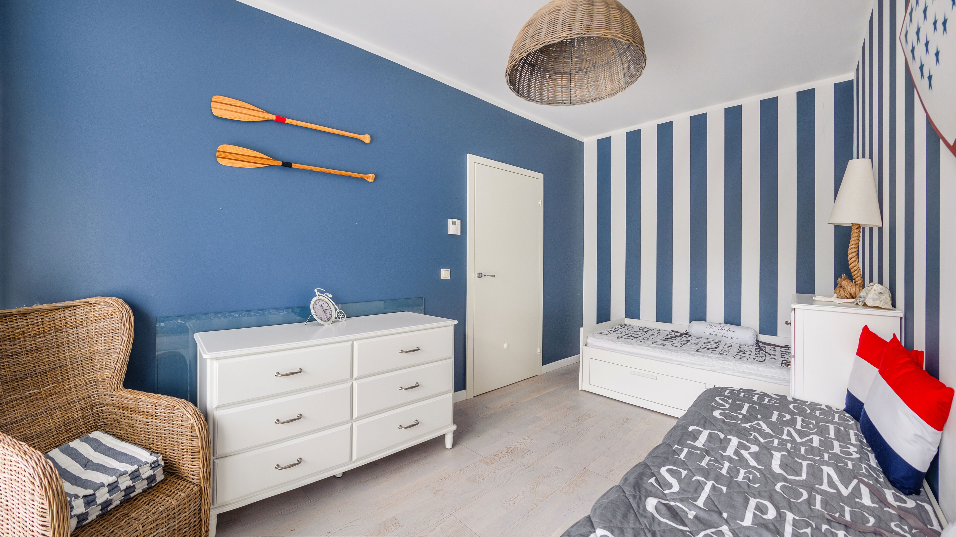 Ein Zimmer mit blauer Wand, einem weißen Schminktisch, einem Bett mit gestreifter Bettdecke und einem Korbstuhl.