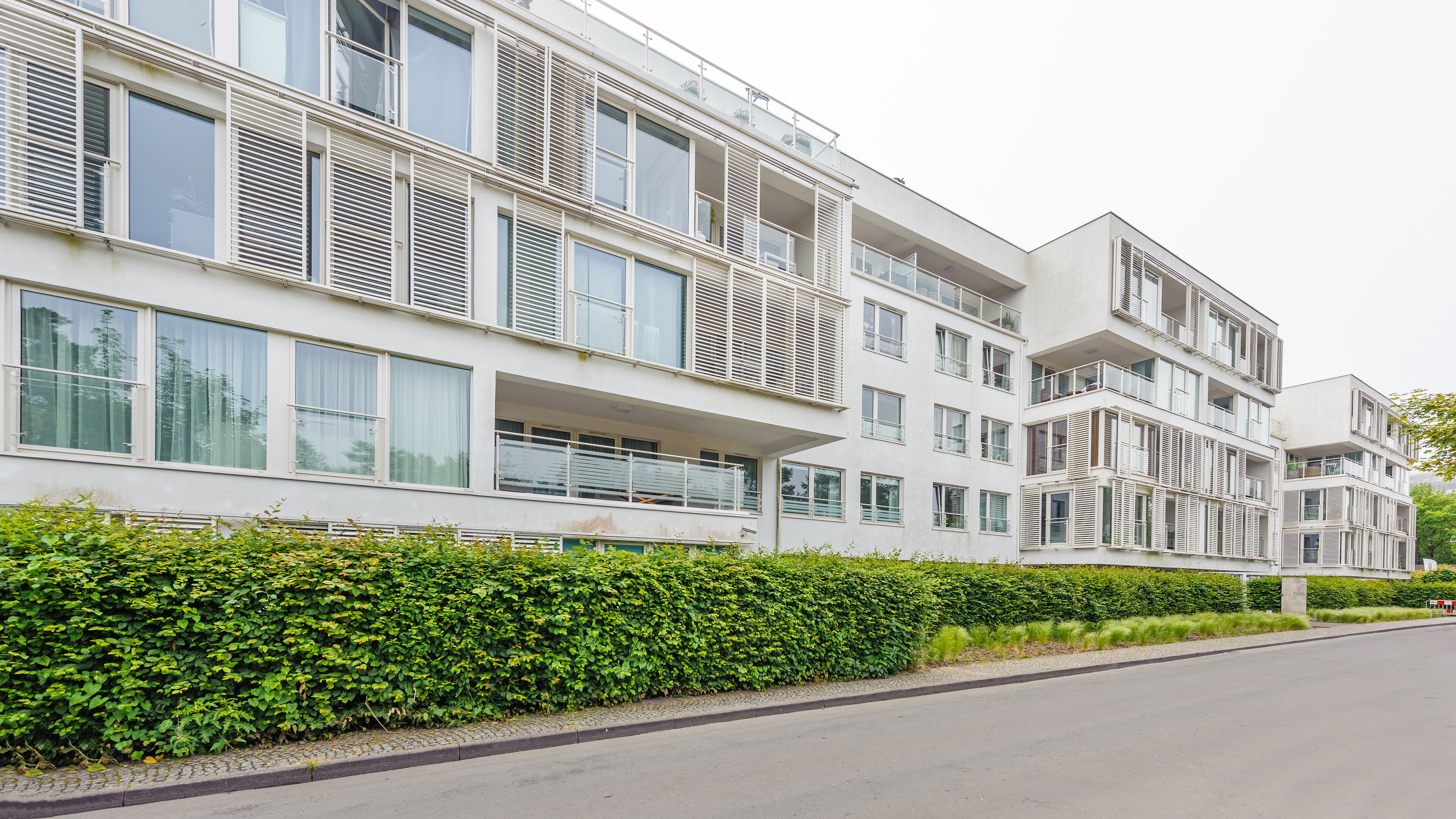 A modern multi-story residential building with balconies, large windows, and a well-maintained hedge along the street.