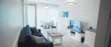 A modern living room with a dark sofa, a small white table, and a TV. There is a dining area with a table and chairs in the background.