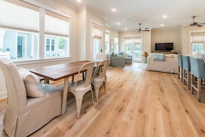 A spacious living area with a wooden dining table, white chairs, a sofa, and a TV.