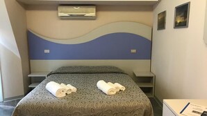 A hotel room with a bed, two rolled towels on the bed, a desk with a chair, an air conditioning unit on the wall, and two framed pictures on the wall.