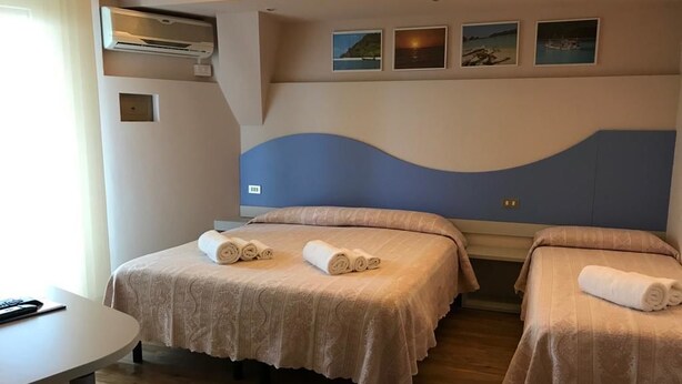 A hotel room with two beds, each with a rolled towel, a desk, and a wall-mounted air conditioner.