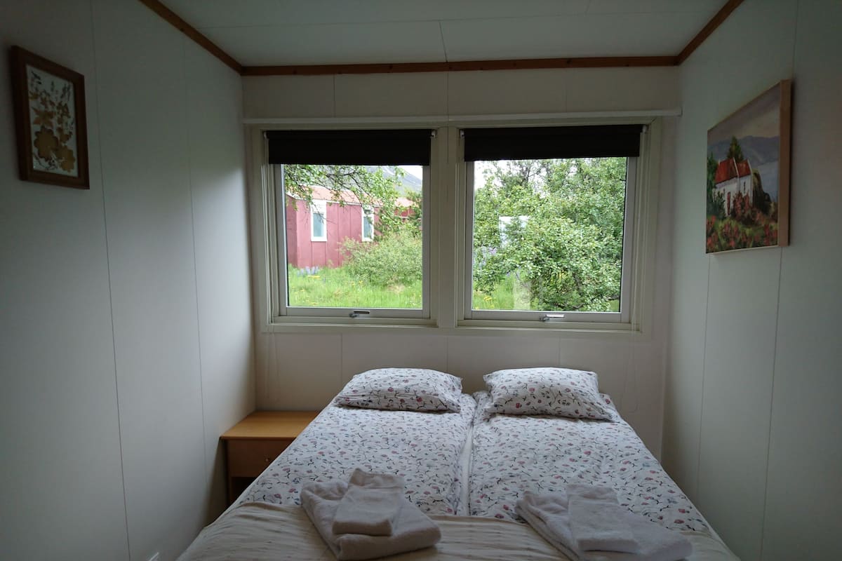 A small, simple bedroom with a bed, two windows, and a view of greenery outside.