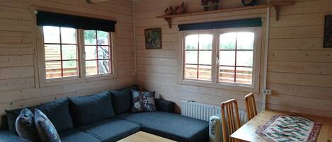 A cozy cabin interior with a dark blue sofa, a small wooden table, and a dining table with chairs. There are two windows with shutters, a radiator, and a shelf with decorative items.