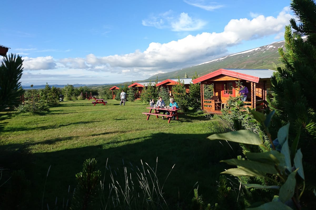 A rustic lodge with wooden cabins, picnic tables, and a grassy landscape with distant mountains.