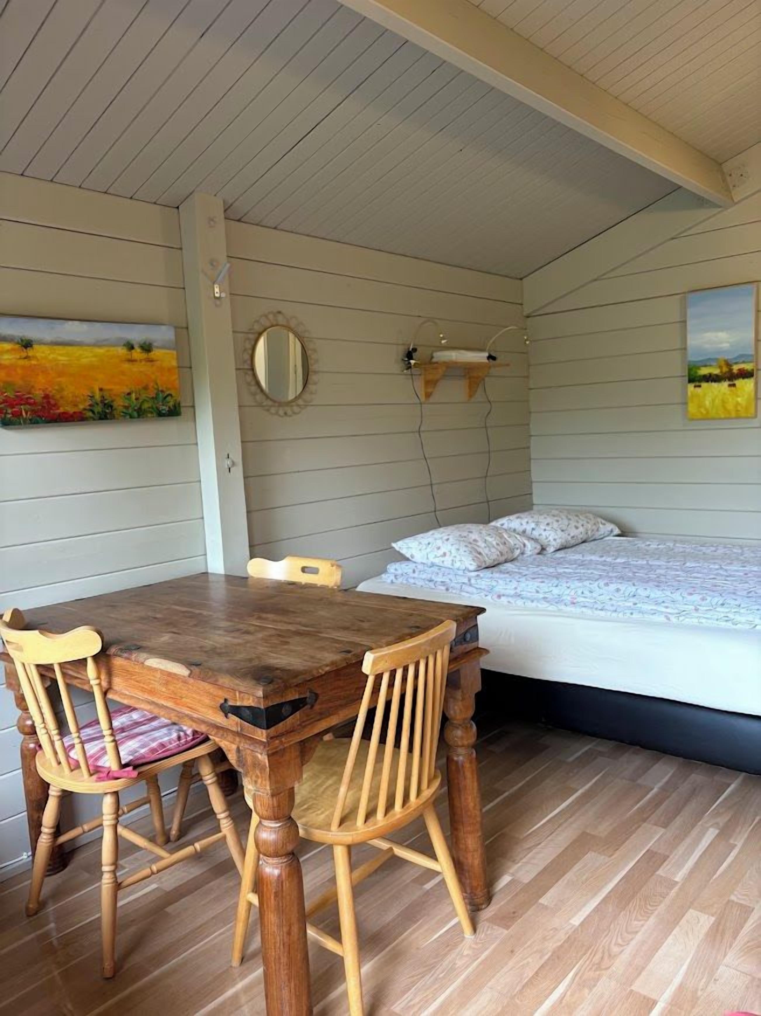 A room with a wooden dining table and chairs, a bed with a white mattress and patterned bedding, and two paintings on the walls.