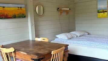 A room with a wooden dining table and chairs, a bed with a white mattress and patterned bedding, and two paintings on the walls.