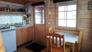 Puinen mökki, jonka sisätiloissa on keittiö, ruokapöytä, tuoleja ja ikkuna.