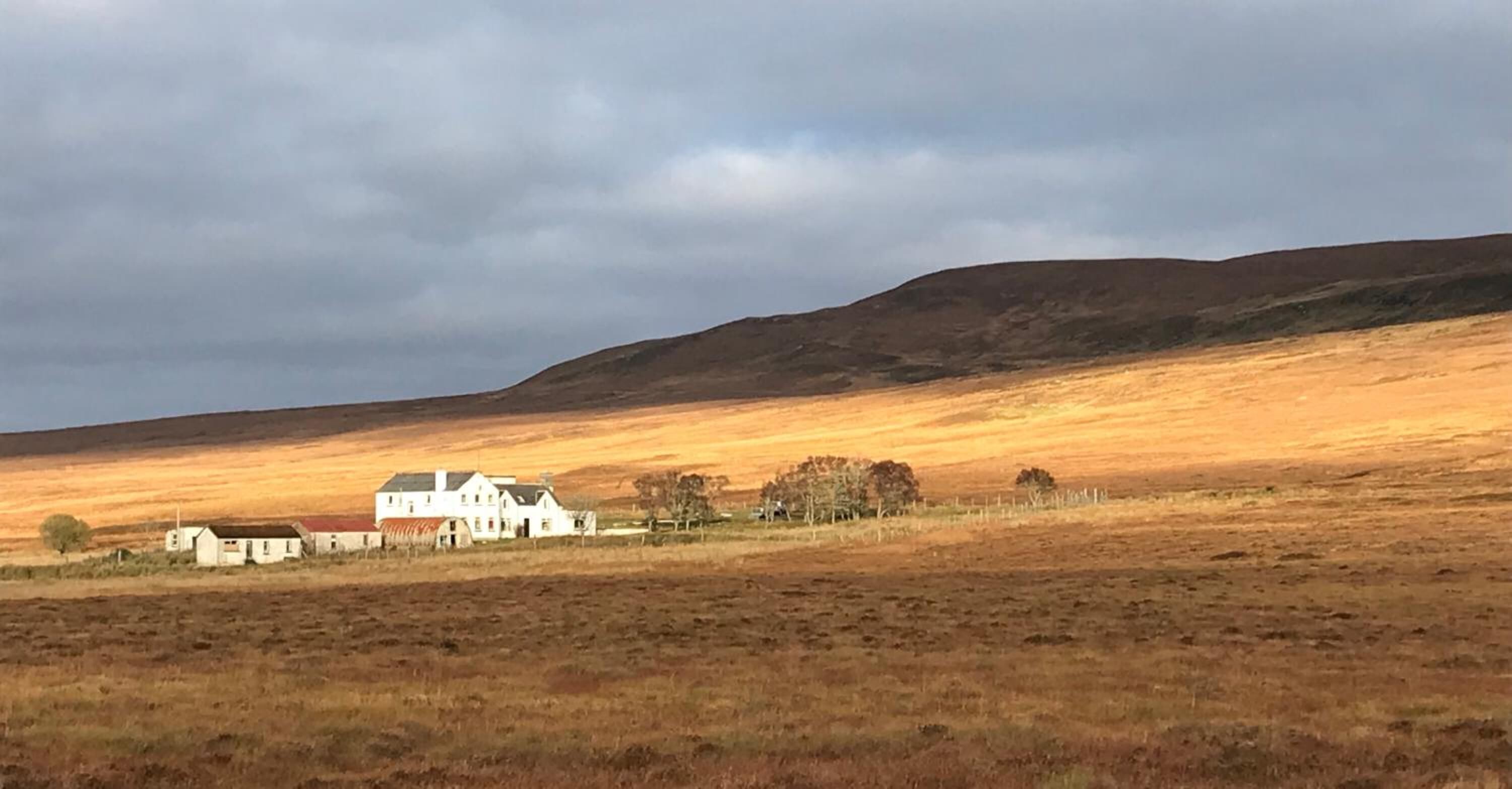 A rural landscape with a white building, possibly a farmhouse, surrounded by open fields and a hill in the background.