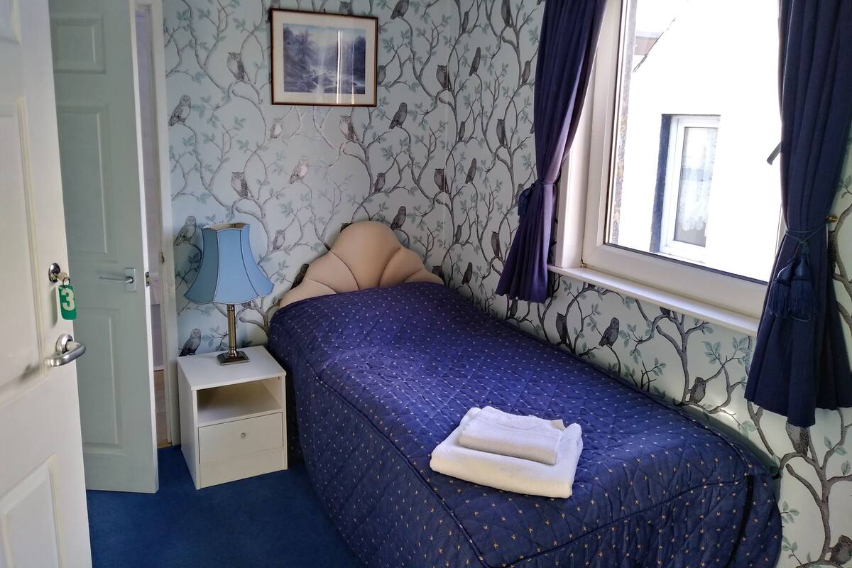 Une chambre à coucher comprenant un lit, une table de chevet, une lampe, une fenêtre avec des rideaux et une porte portant un numéro.