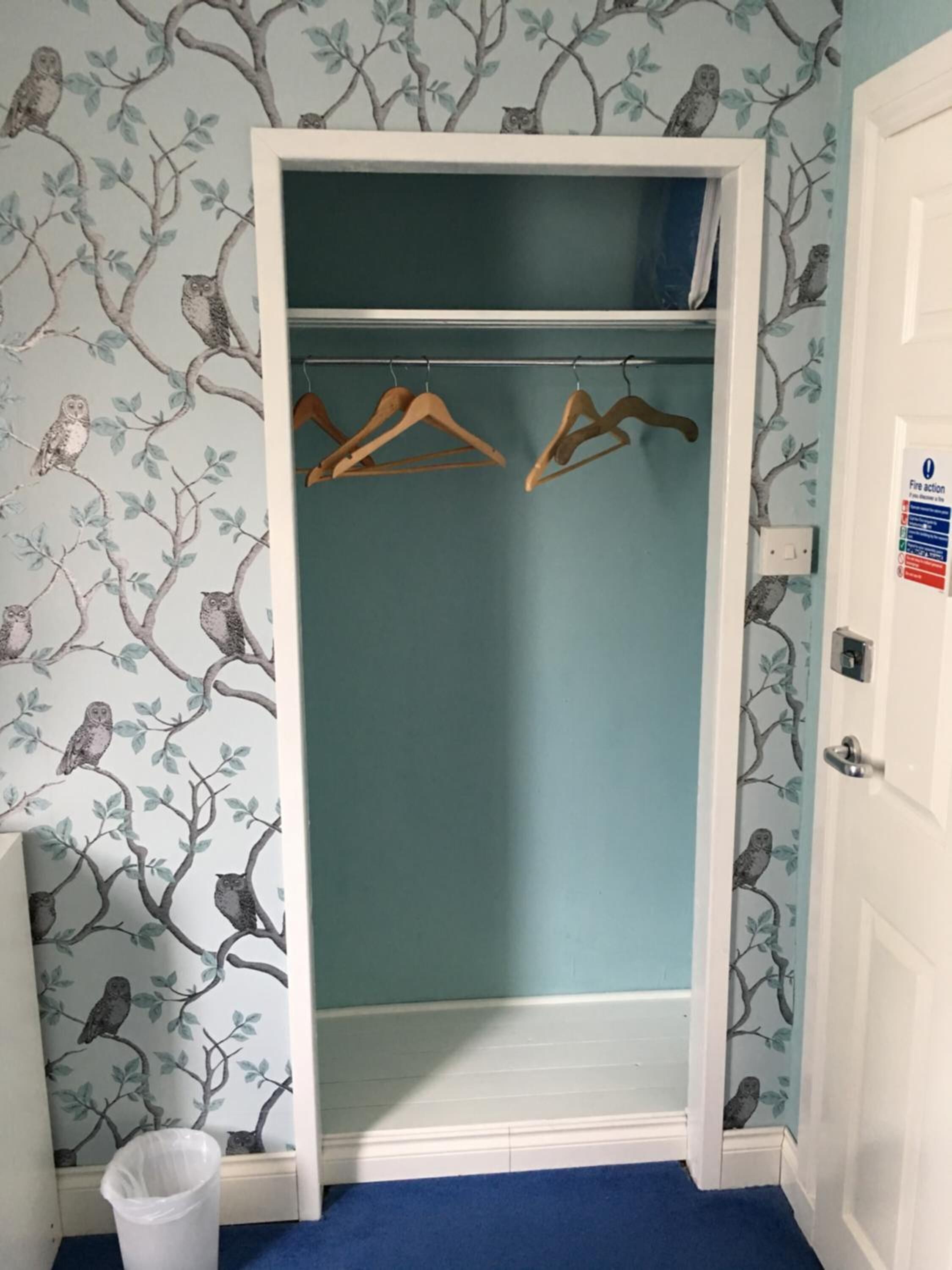 Vestidor con pared azul y papel tapiz con ramas y lechuzas. Se ven perchas de madera en una barra y un bote de basura blanco a la izquierda.