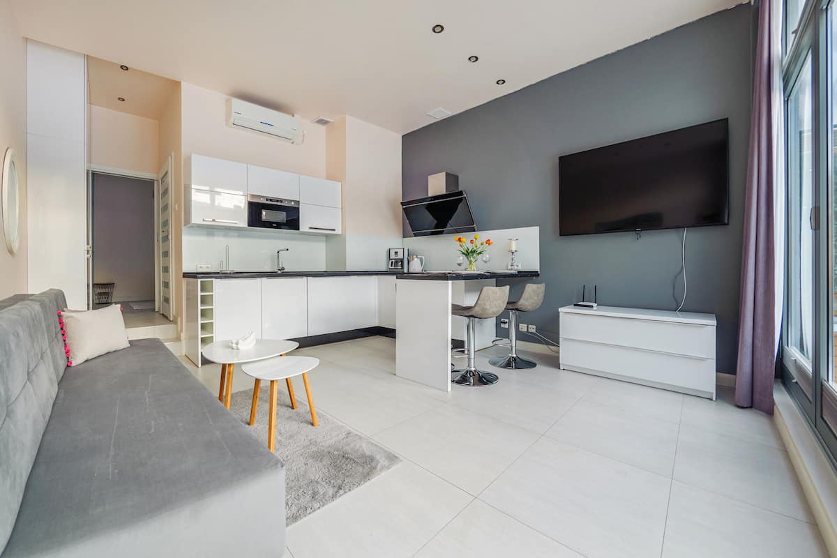 A modern kitchen with white cabinets, a black countertop, and a built-in microwave. There is a dining area with two chairs and a small table. A grey sofa is in the living area, and a flat-screen TV is mounted on the wall.