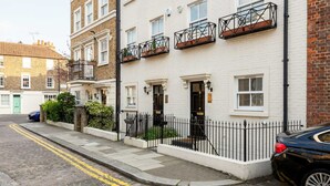 Une rangée de maisons de ville aux façades en briques blanches, dotées de balcons avec des garde-corps noirs, une rue pavée et une voiture garée.