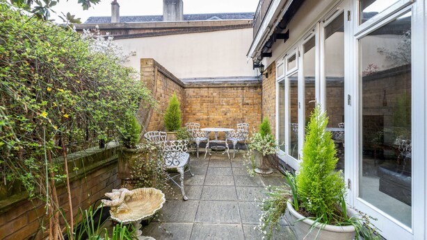 Une terrasse avec un mur en briques, une clôture en bois, ainsi qu’une table et des chaises en métal peintes en blanc.