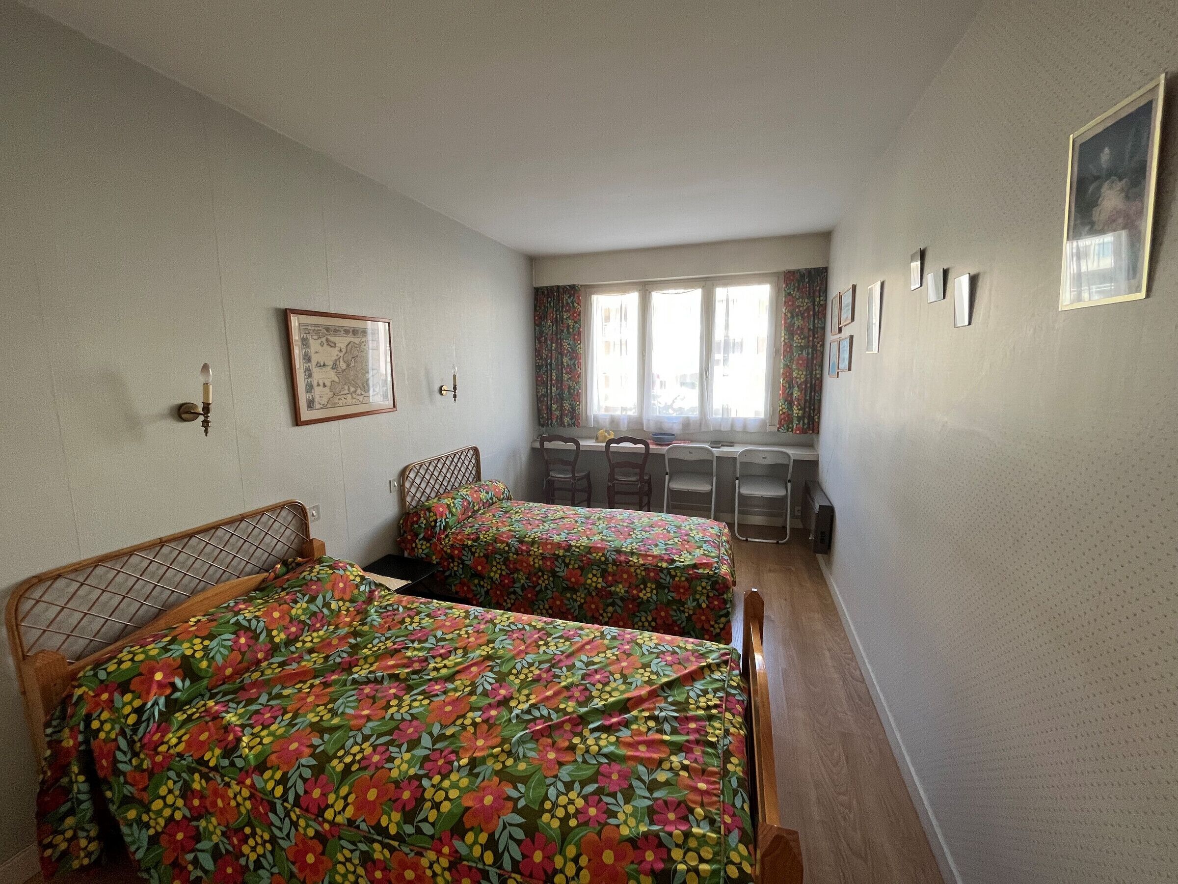Habitación con dos camas, un escritorio y una silla. Ventanas con cortinas, una lámpara de pared y cuadros enmarcados.