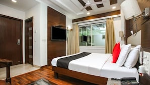 A hotel room with a wooden floor, a bed with white and black bedding, a TV mounted on the wall, and a window with curtains. - West Yard Hotels (New Delhi)