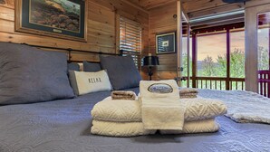 A wooden cabin room with a bed, pillows, a framed picture, and a window with a view.