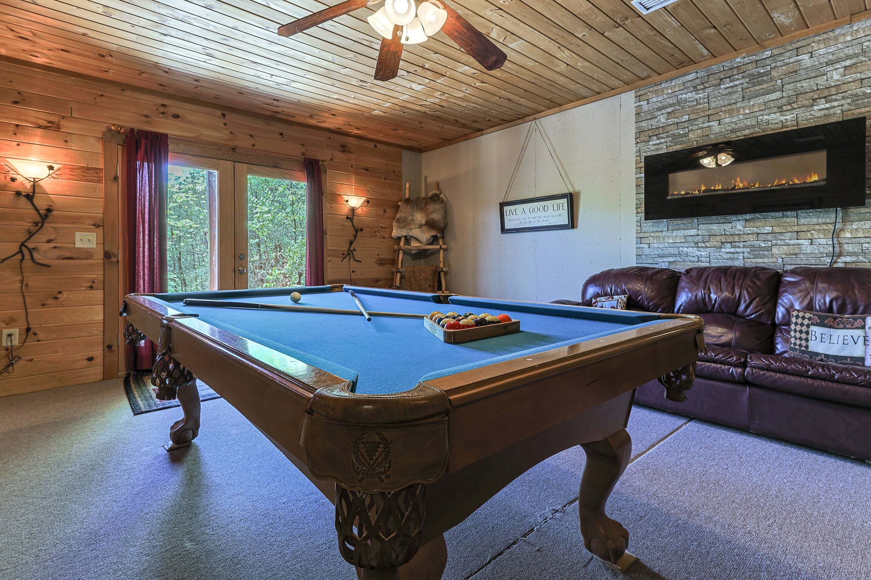 A pool table in a room with wooden walls, a stone fireplace, and a leather couch.