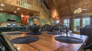 A wooden cabin interior with a dining table, chairs, a kitchen area, and a staircase.