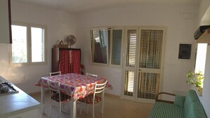Un comedor con una mesa cubierta por un mantel estampado, sillas blancas y un ventilador. Se observa un sofá verde y una ventana con cortinas al fondo.