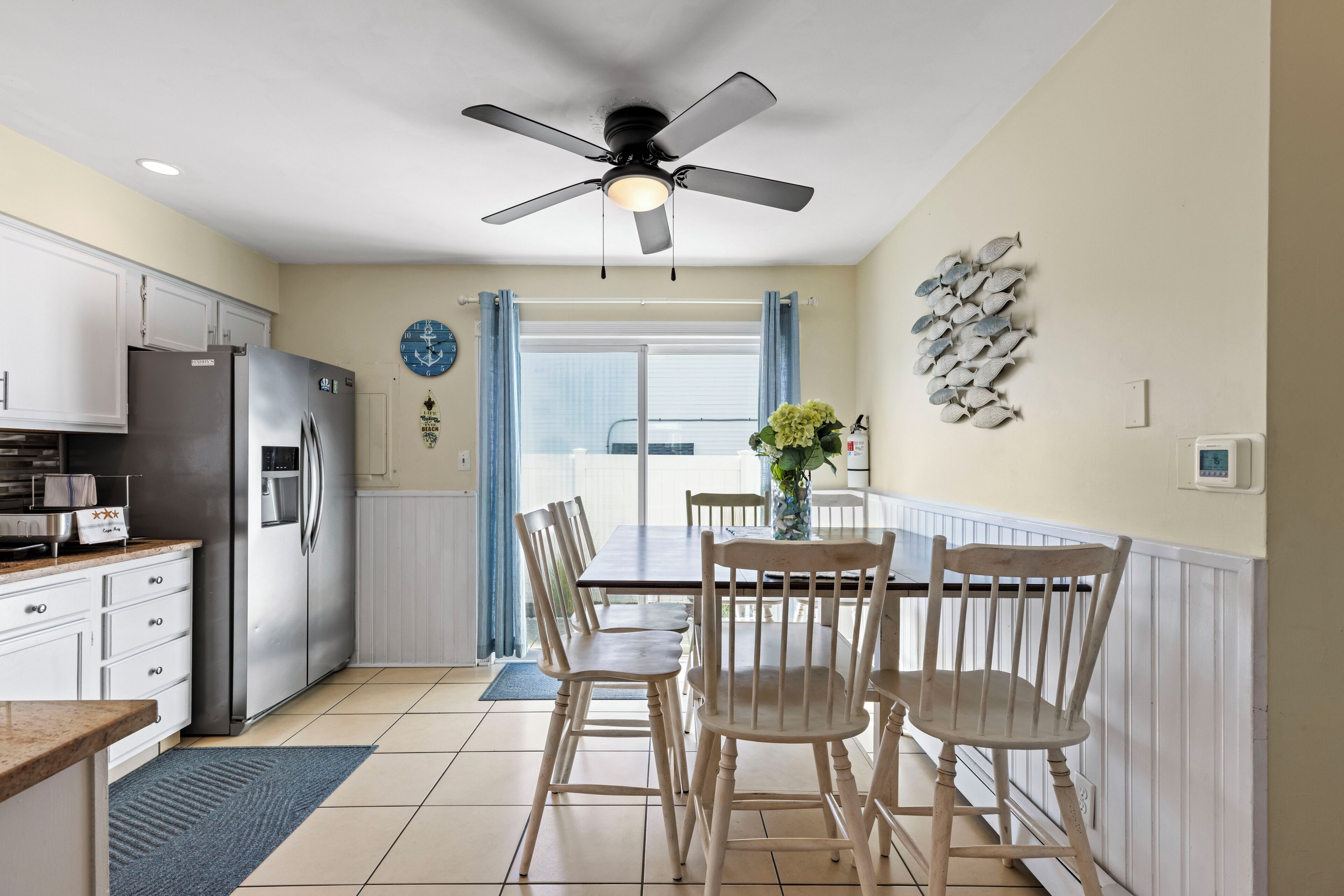 A kitchen with white cabinets, a stainless steel refrigerator, a dining table with chairs, a ceiling fan, and a window with curtains.