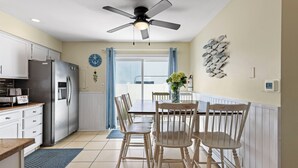 Una cucina con mobili bianchi, un frigorifero in acciaio inox, un tavolo da pranzo con sedie, una ventola a soffitto e una finestra con tende.