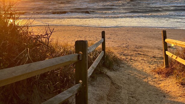 Una spiaggia con una recinzione di legno che conduce alla riva, spiaggia sabbiosa e onde oceaniche sotto un cielo al tramonto.