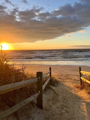 Une scène de plage avec une clôture en bois menant au rivage, une plage de sable et des vagues déferlant sur l’océan, le tout sous un ciel au coucher du soleil.