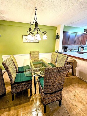 A dining area with wicker chairs and a glass table, green walls, and a kitchen in the background.