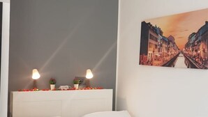 Een hotelkamer met een bed, twee bedlampen, een decoratieve plank met bloemen, en een stadsgezicht schilderij aan de muur.